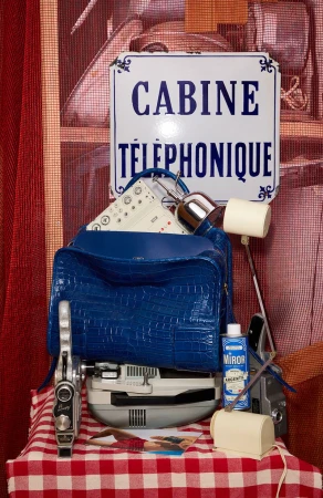 Retro display featuring a blue handbag, vintage cameras, cleaning supplies, and a French telephone booth sign on a checkered tablecloth.