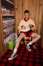 Model wearing yellow sweater and red shorts holding a detergent bottle in a wood-paneled room with grocery shelving
