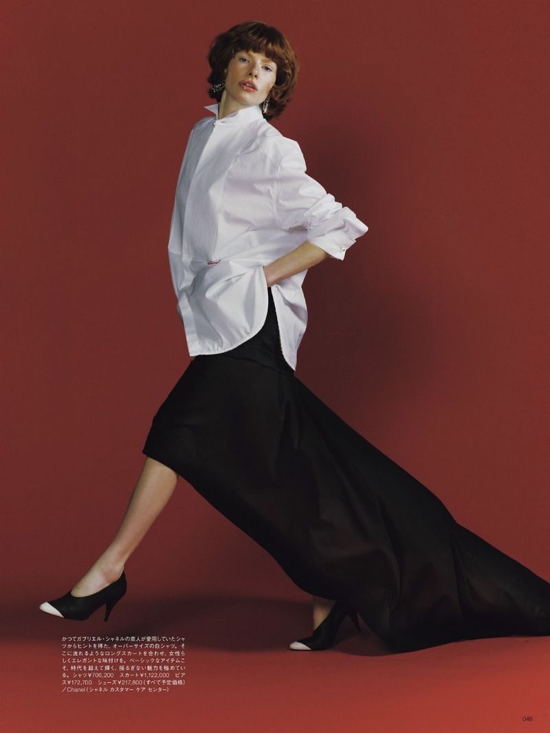 Fashion editorial shot of a woman in a white shirt and long black skirt striking a pose against a solid red backdrop with studio lighting.