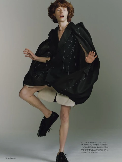 Fashion shot of a model in oversized blazer and black shoes with fringe, standing on one leg against a solid grey backdrop in studio light.