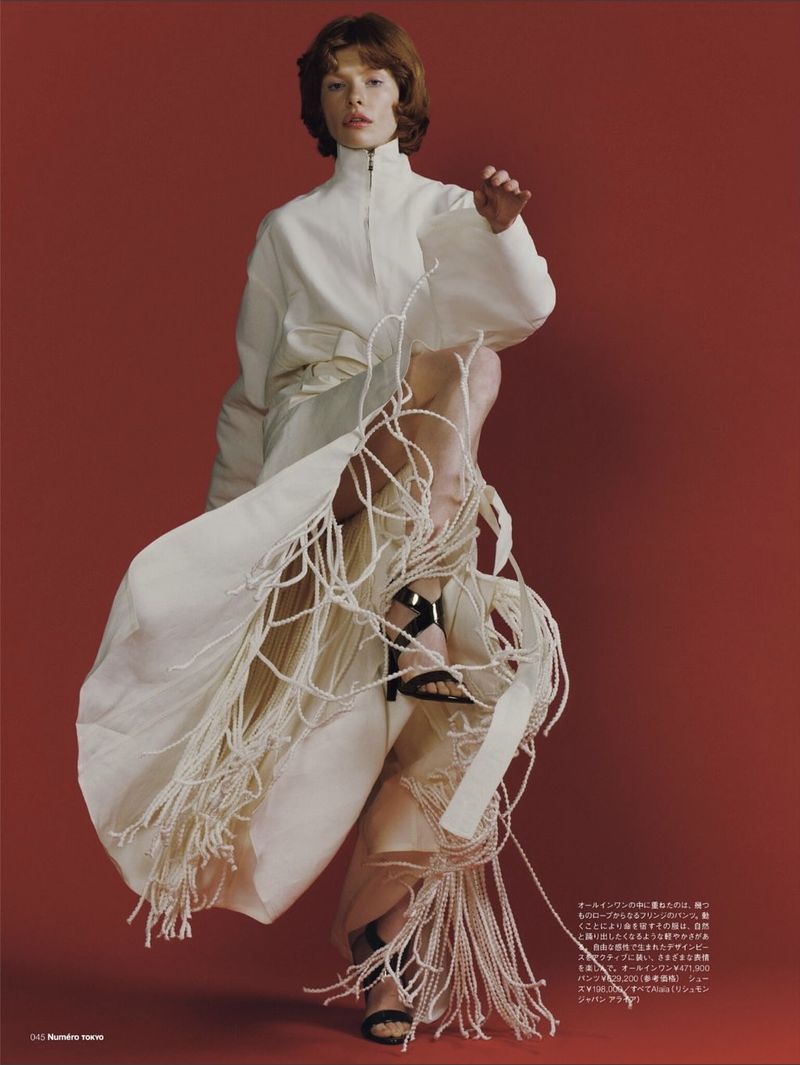Fashion editorial shot: A young woman in white dress and black heels poses against a red background in a full-length studio shot.