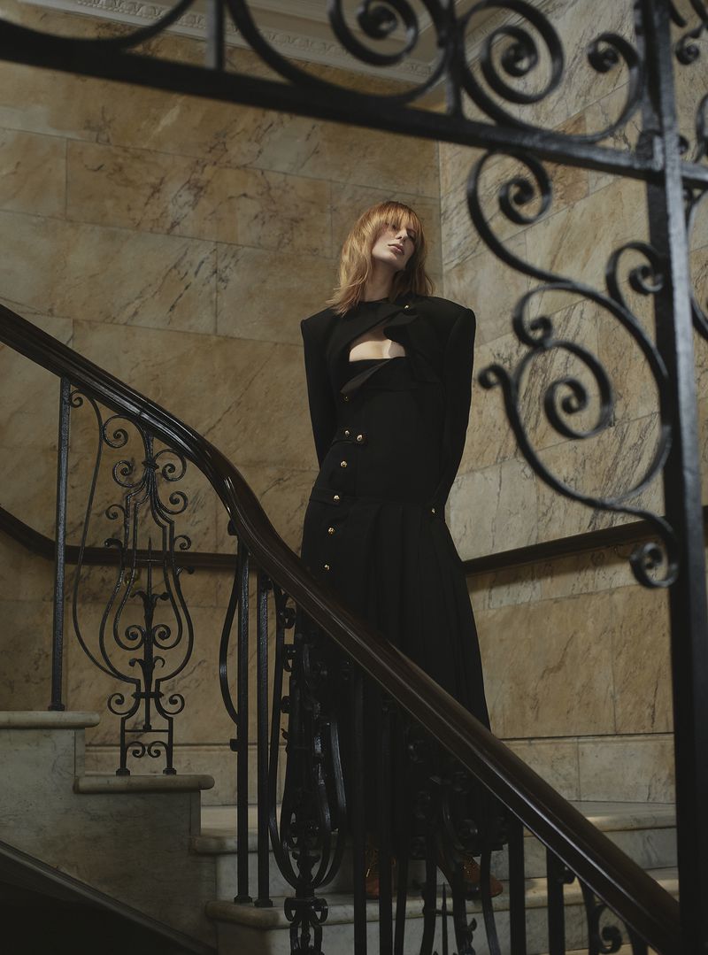 Woman posing on stairs in fashionable black cut-out dress with shoulder pads and gold buttons indoors against beige marble wall.