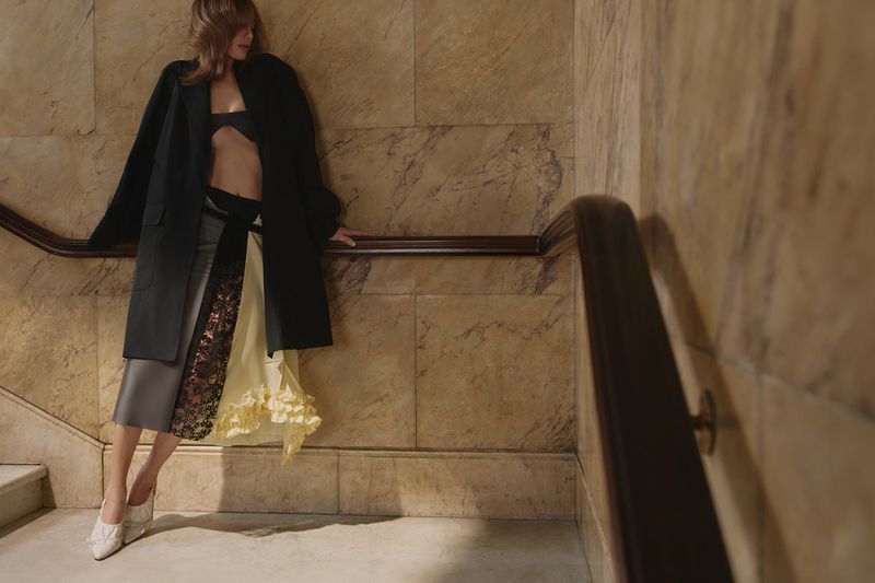 An adult woman in high fashion poses on a staircase. She wears an open black coat, bra top, and layered skirt. The background is a stone wall.