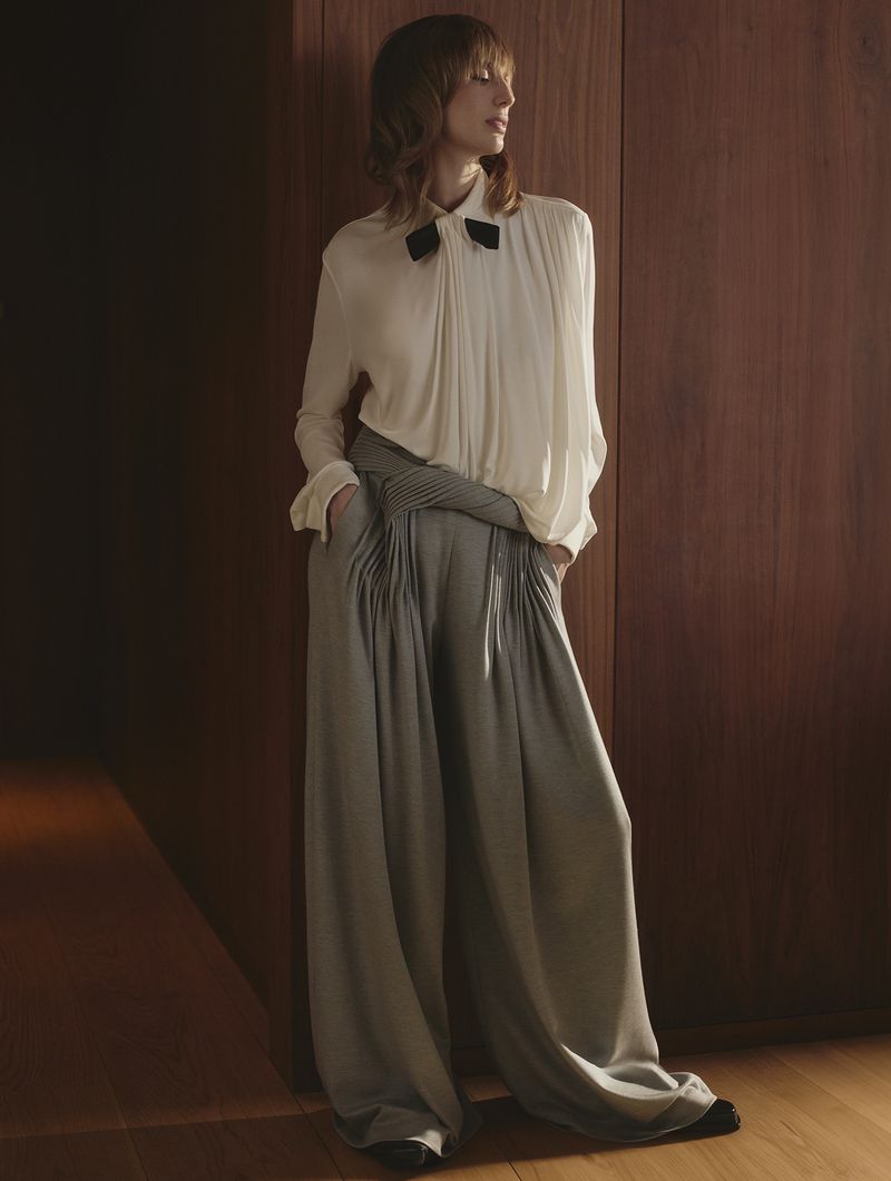 Stylish woman in white blouse and grey palazzo pants posing in front of dark wood paneling, fashion photography, studio lighting.