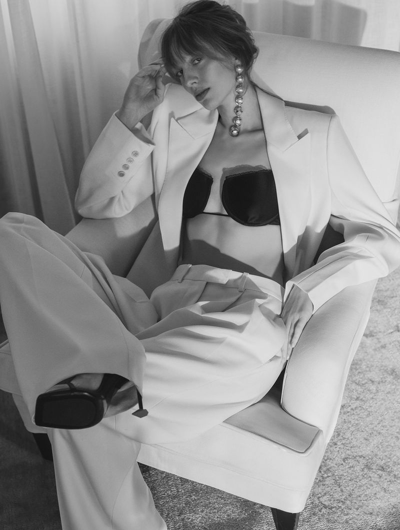Elegant woman in a white suit and black bra top poses in a white armchair with large pearl earrings in a monochrome fashion shot.