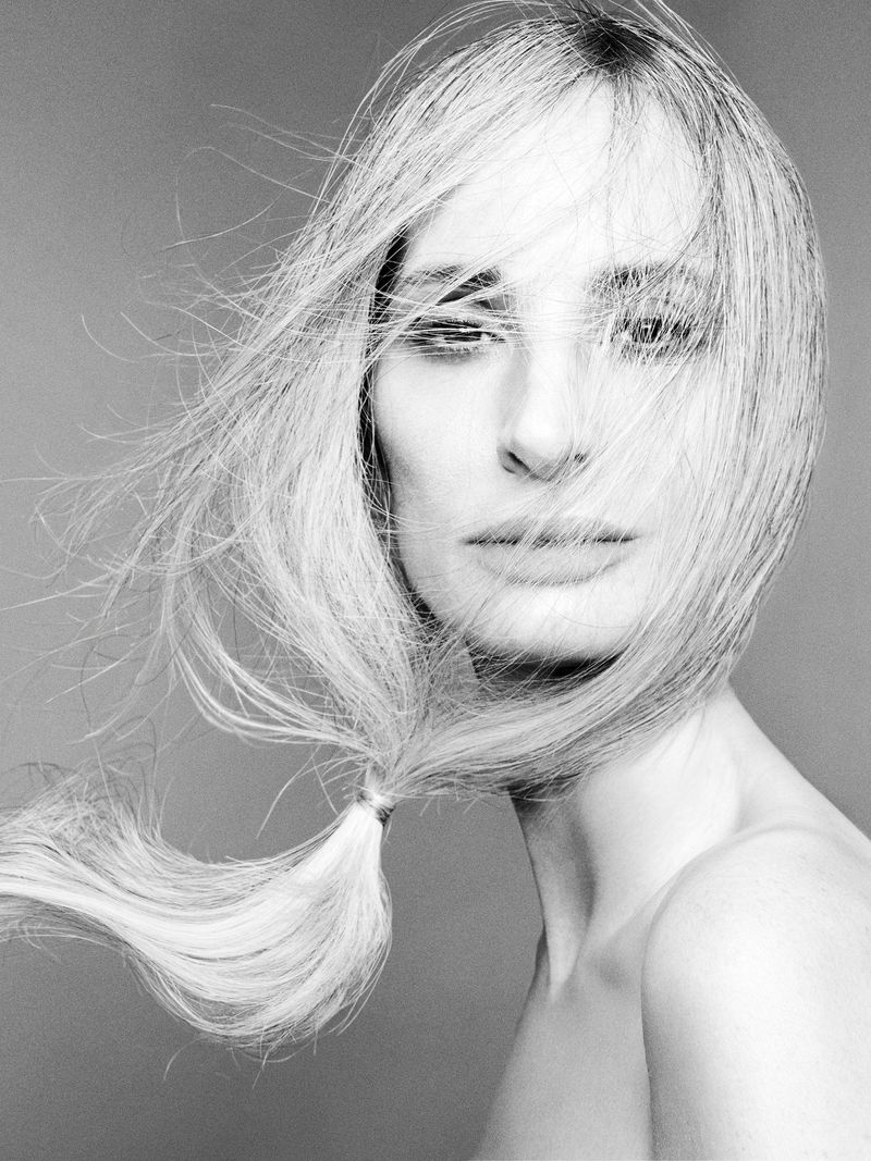 Close-up portrait of a woman with long hair blowing across her face against a plain grey background