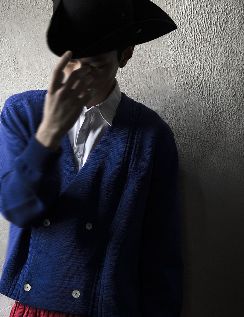 Man poses in a dark photo studio wearing a black hat and blue cardigan, with red pants visible. A textured wall is behind him.
