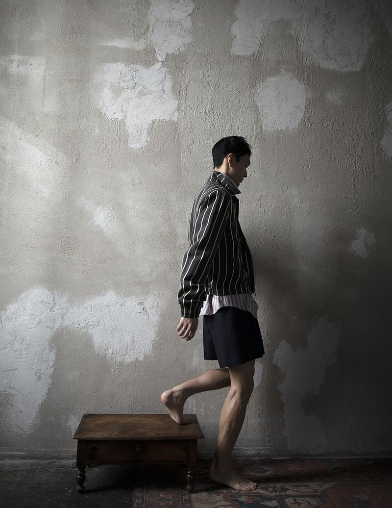A slender man wearing a striped jacket and shorts steps onto a wooden stool indoors against a textured grey wall, barefoot with a vintage rug.