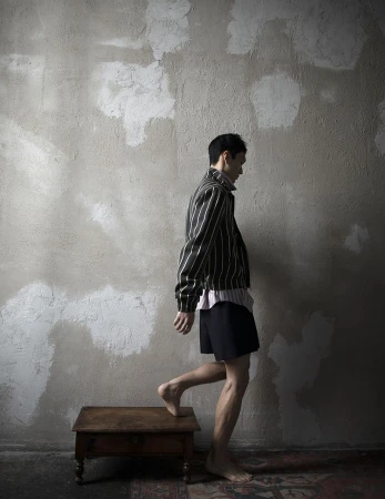 A slender man wearing a striped jacket and shorts steps onto a wooden stool indoors against a textured grey wall, barefoot with a vintage rug.