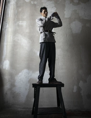 A standing Asian man in a gray outfit and patterned pants is striking a pose atop a black step stool in an indoor studio setting.