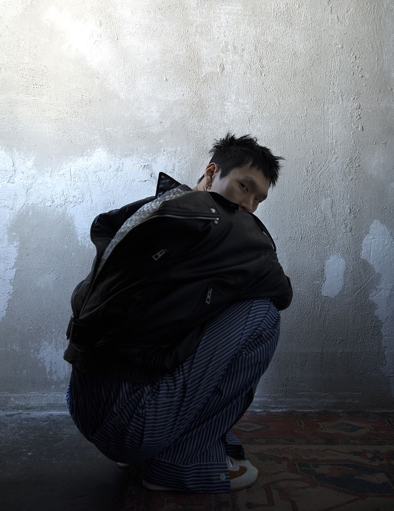 Asian man in black jacket and striped pants squatting in a room with concrete wall and patterned rug, looking at the camera with a serious expression.