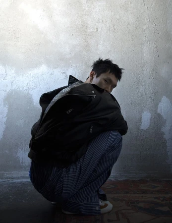 Asian man in black jacket and striped pants squatting in a room with concrete wall and patterned rug, looking at the camera with a serious expression.