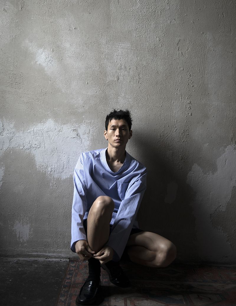 Asian man in a blue shirt is crouching against a textured grey wall indoors. He is wearing black socks and shiny black shoes.