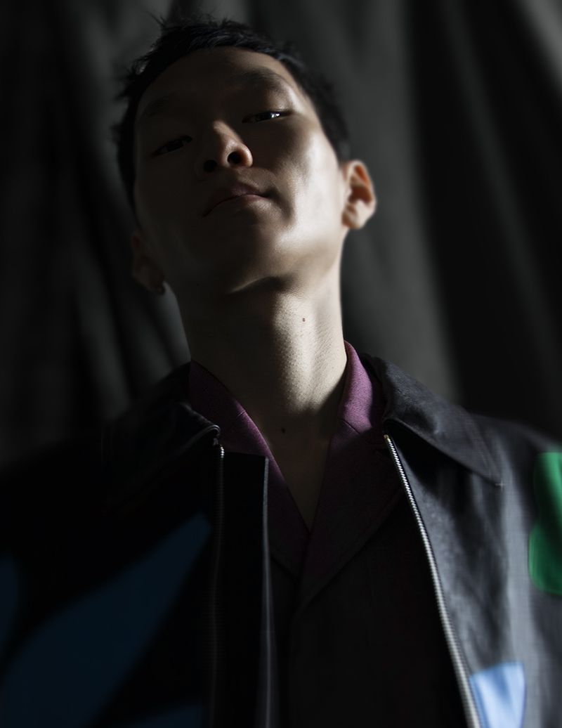 Close-up studio shot of a young adult male in a black jacket with a purple collared shirt, under dark lighting, against a black backdrop.
