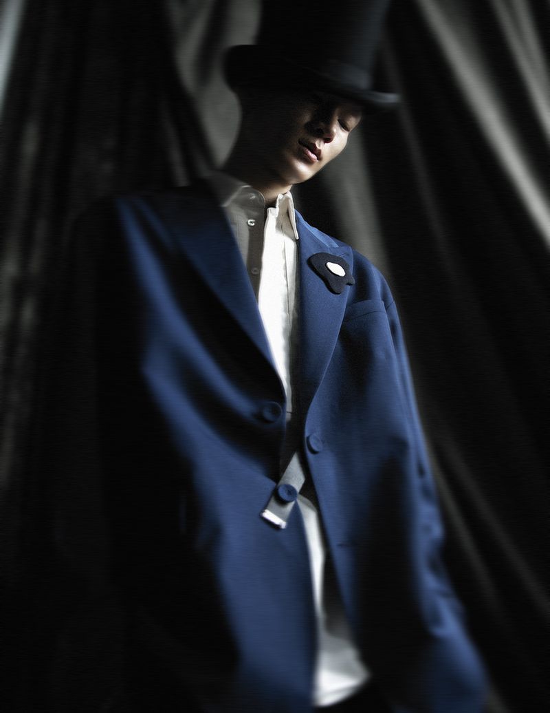 Man in a formal blue suit and top hat poses dramatically in a dimly lit setting with dark, draped fabric in the background. Fashion shoot.