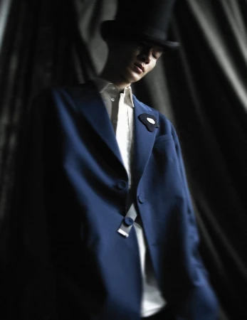 Man in a formal blue suit and top hat poses dramatically in a dimly lit setting with dark, draped fabric in the background. Fashion shoot.