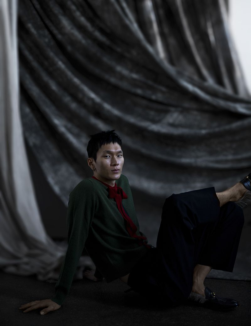 Portrait of an Asian male model in a dark green sweater and black pants, sitting against a draped, dark gray fabric backdrop.