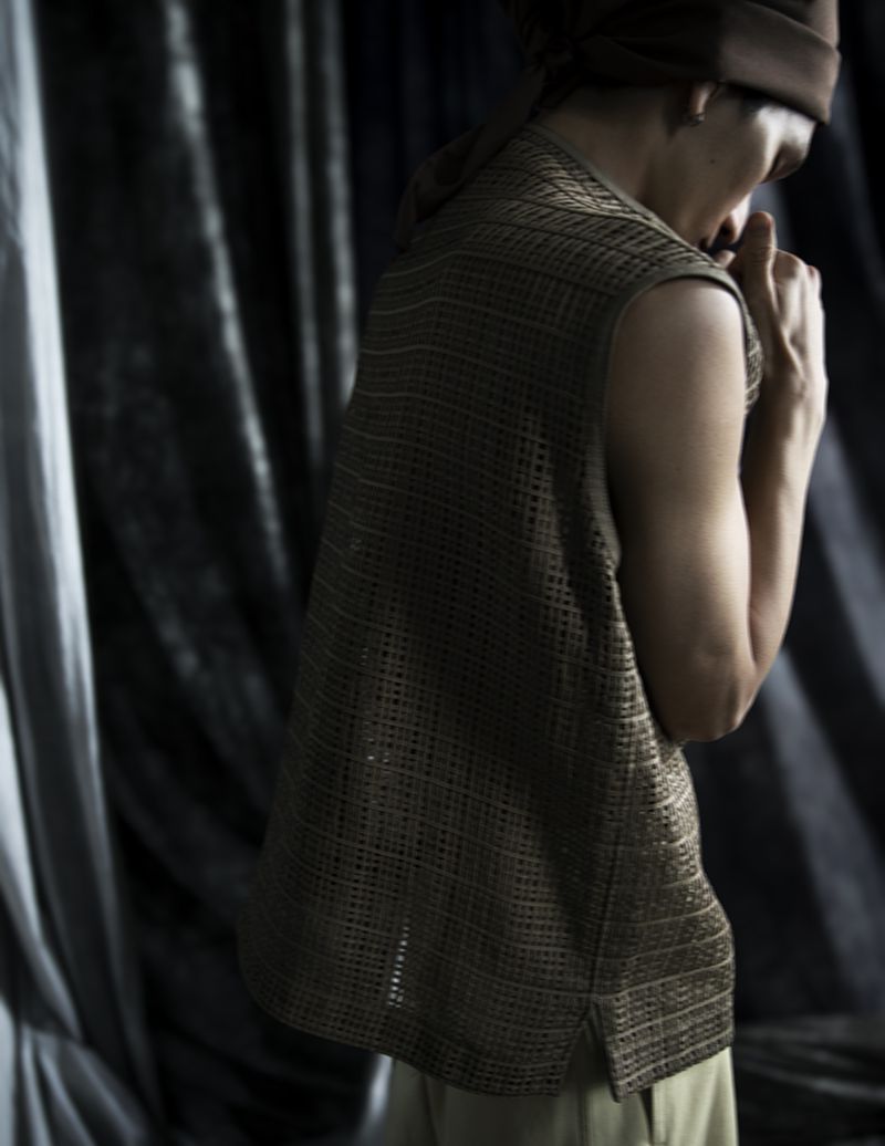 Person in a brown knit sleeveless shirt and hat stands against a draped fabric backdrop in a studio setting with soft, diffused light.