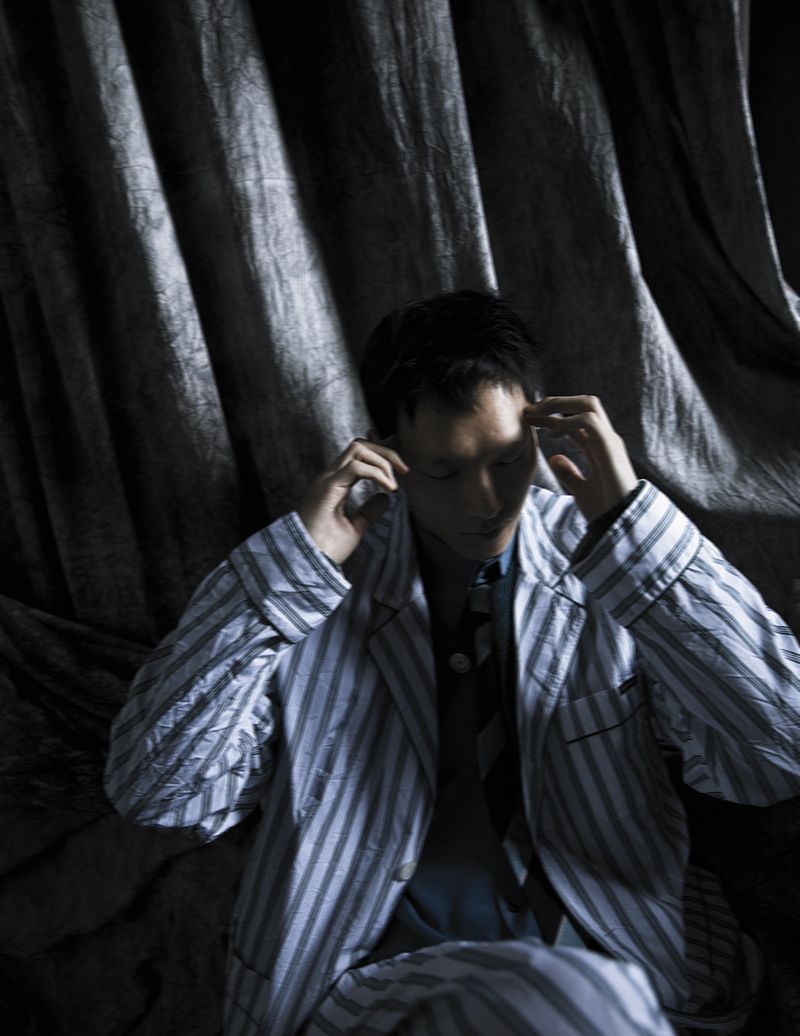 Adult male in striped pajamas with hands on his head, indoor shot against a dark, draped background, creating an intimate and moody portrait.