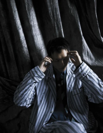 Adult male in striped pajamas with hands on his head, indoor shot against a dark, draped background, creating an intimate and moody portrait.