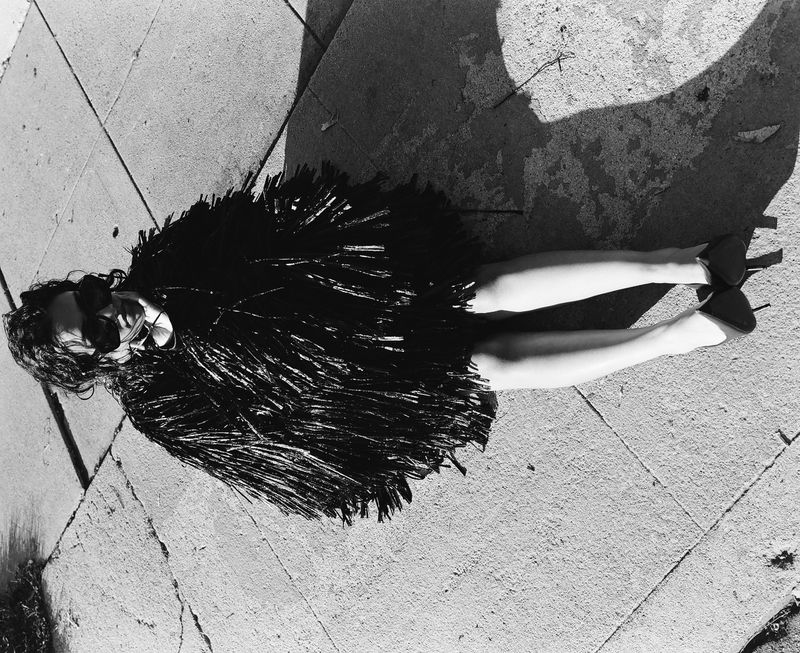 Overhead shot of a woman lying on a stone surface wearing a fringed jacket, sunglasses, and high heels, creating a striking fashion moment.