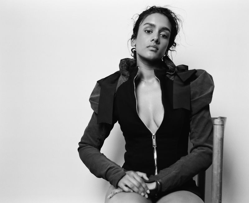 Black and white portrait of a young woman seated on a chair, wearing a plunging-neckline bodysuit with a jacket featuring statement shoulders.