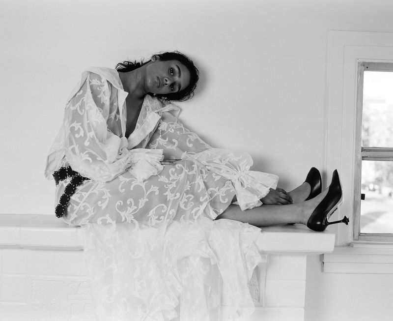 A woman wearing a patterned white lace blouse and matching skirt sits on a white ledge in a monochrome portrait with natural window light.