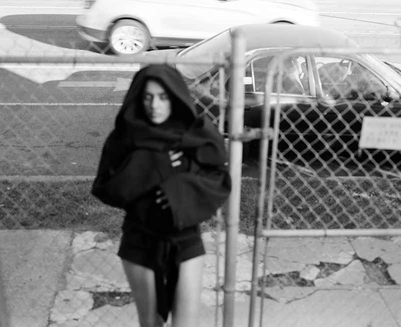 Adult female wearing a hoodie and shorts is the primary focus, with cars visible in the blurry background, presented in black and white.