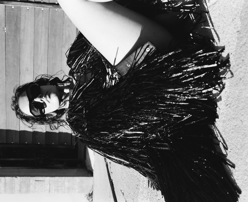 A stylish woman in sunglasses and a fringed top poses outdoors in a black and white image, creating a high-contrast fashion statement.