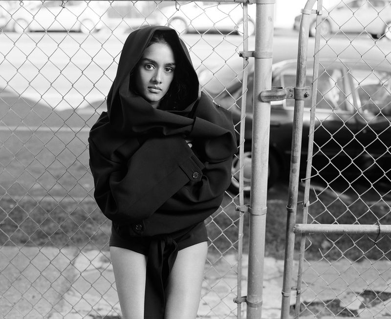 A woman in a black outfit stands in front of a chain-link fence, creating a striking portrait with urban background details and monochrome aesthetic.
