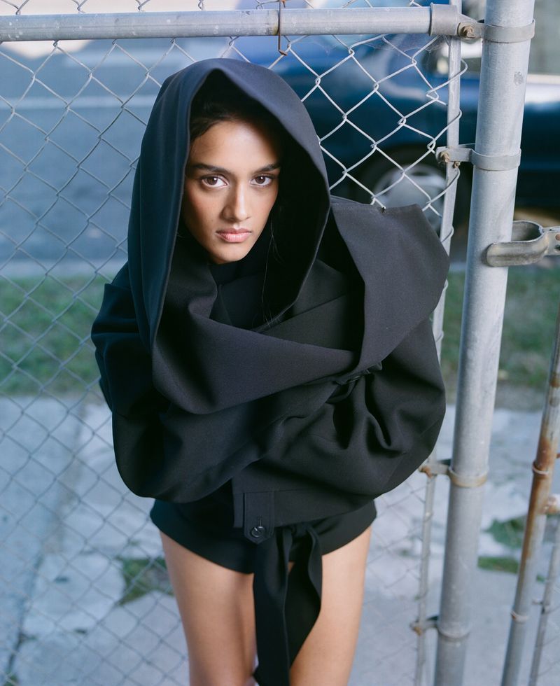 Woman in dramatic black hooded jacket poses in front of gray chain-link fence. Fashion editorial shot from a low angle. 
