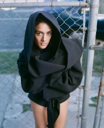 Woman wearing a black hooded garment standing against a chain-link fence for the Shabana Azeez project by Side-note.