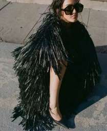 A woman wearing a black fringed coat and large sunglasses seated outdoors on pavement.