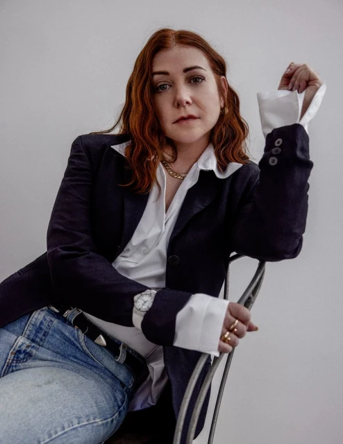 Portrait of an adult woman with auburn hair, seated in a studio wearing a navy blazer, white shirt, jeans, and jewelry.