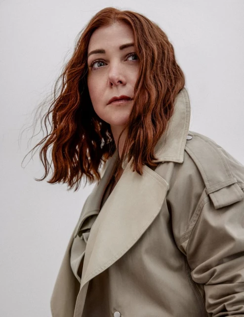 Red-haired adult woman in a beige trench coat looking up and to the side against a neutral backdrop. Shoulder-length wavy hairstyle.