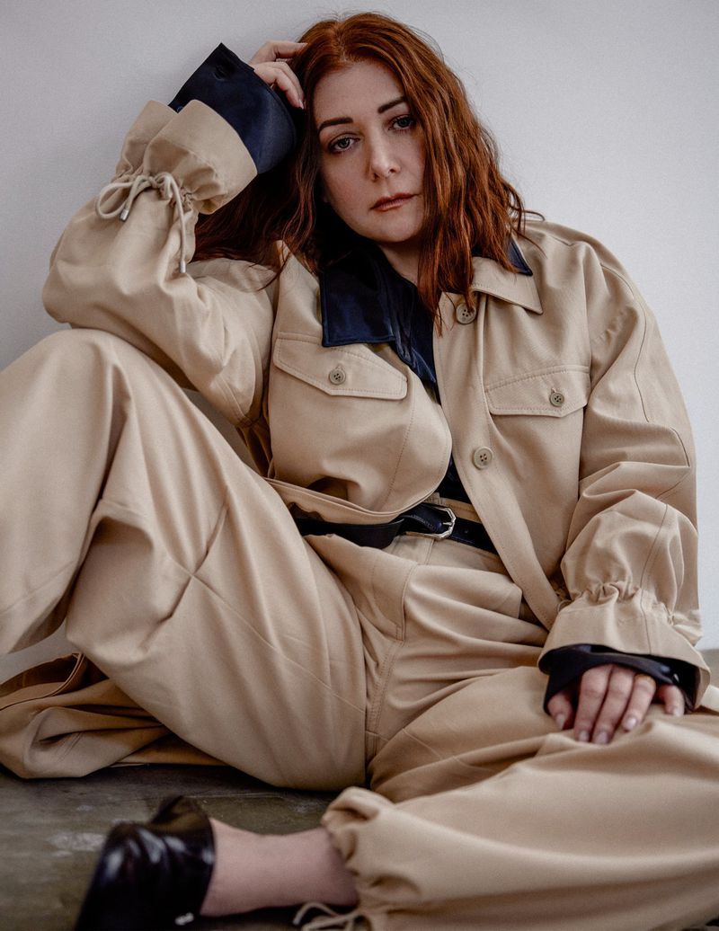 A red-haired adult female fashion model wearing a beige jacket and matching pants is seated against a white wall in a portrait style.