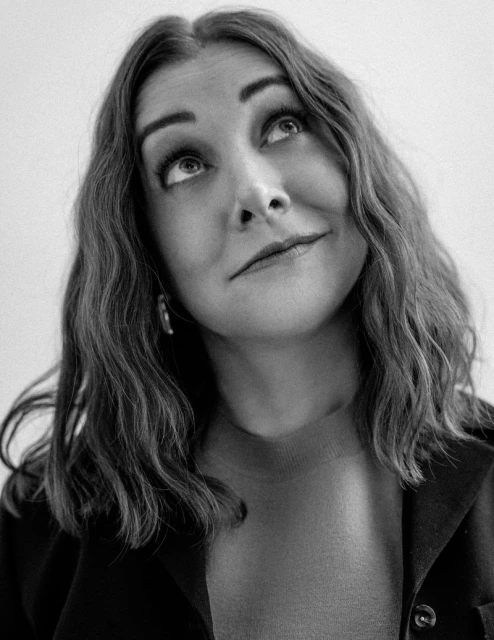 Black and white portrait of an adult female looking up, wearing a blazer and sweater. Studio lighting and a neutral background.