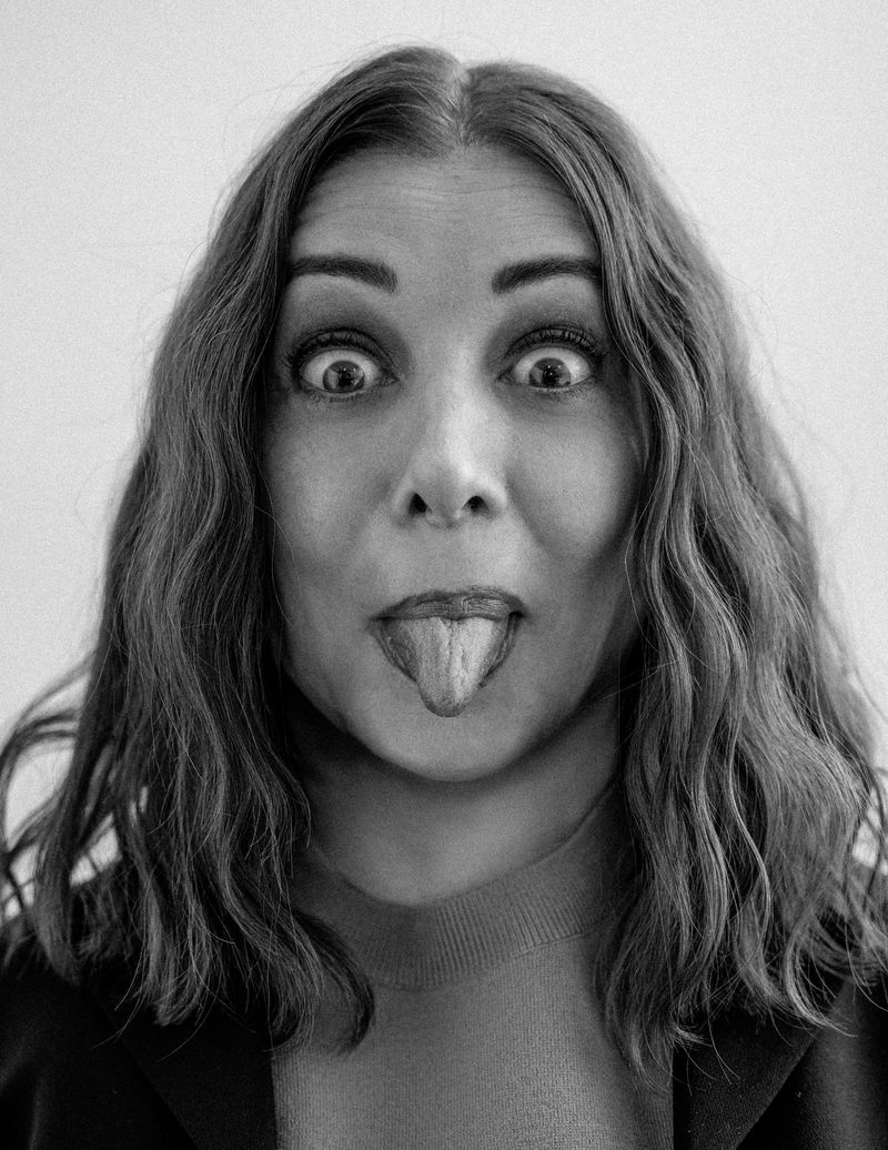 Portrait of an adult female with shoulder-length brunette hair, sticking her tongue out, and pulling a funny face against a white backdrop.