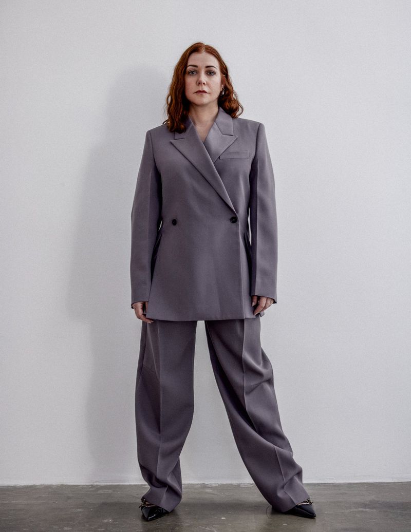 Elegant woman in a grey suit poses against a plain white wall, showcasing fashion and style in a studio setting with clean aesthetics.