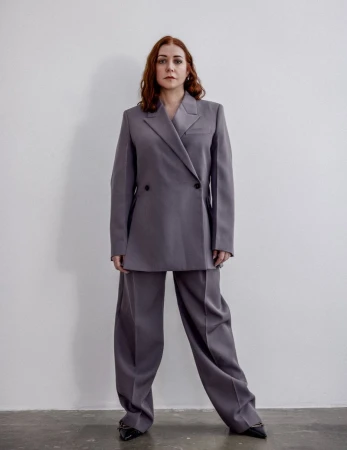 Elegant woman in a grey suit poses against a plain white wall, showcasing fashion and style in a studio setting with clean aesthetics.