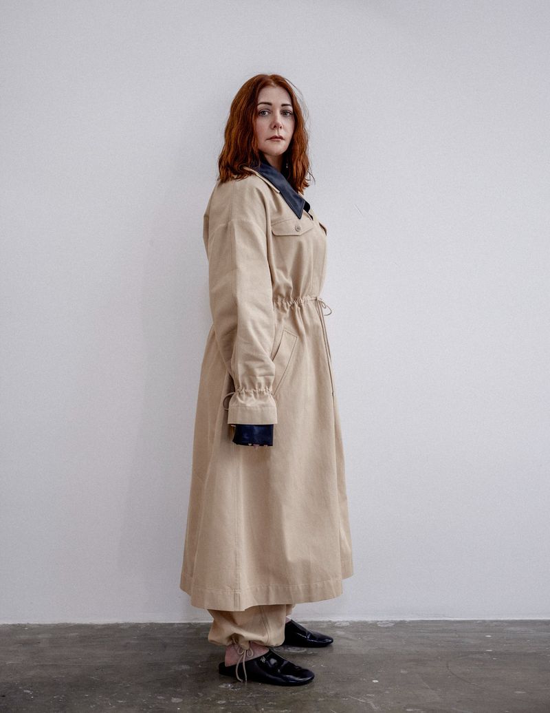 Standing woman with red hair poses in a beige trench coat and trousers against a white wall in a full shot fashion photograph.
