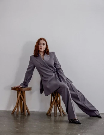 A poised red-haired woman in a grey pantsuit reclines elegantly on a wooden stool against a plain white backdrop in a fashion editorial.