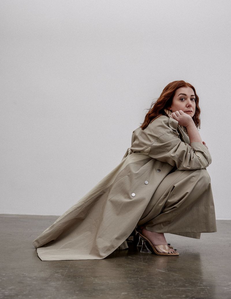 Portrait of a red-haired woman in a beige trench coat, crouching on a gray floor against a white wall, captured in an eye-level shot.