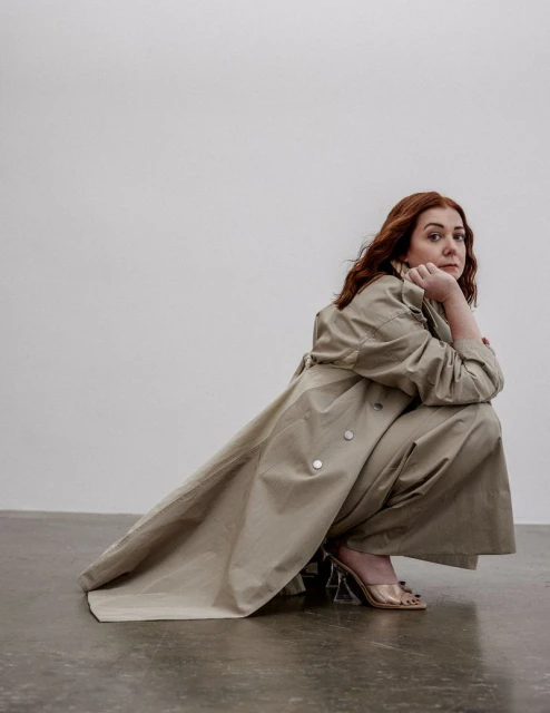 Portrait of a red-haired woman in a beige trench coat, crouching on a gray floor against a white wall, captured in an eye-level shot.