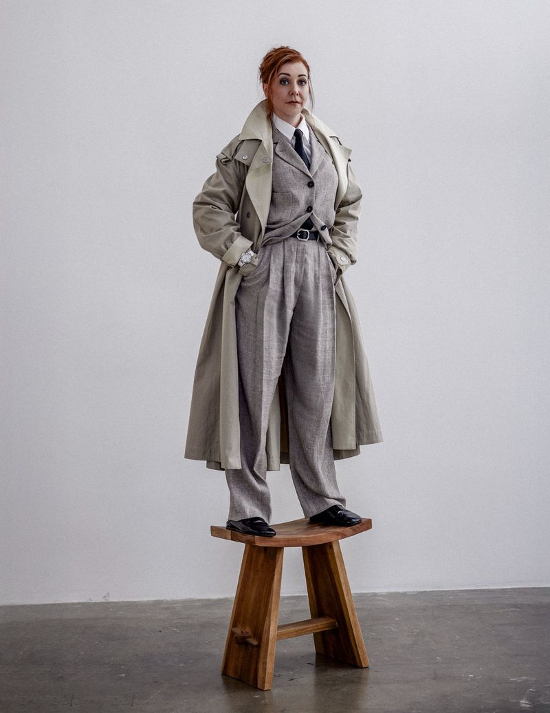 Full shot of a red-haired woman in a grey suit and trench coat standing on a wooden stool in a photo studio with a white background.