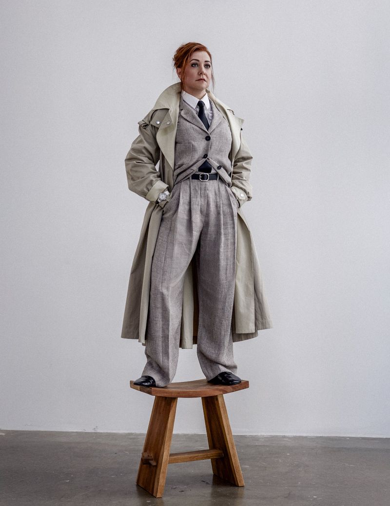 Woman in a gray suit and tan trench coat standing on a wooden stool in a minimalist setting, looking upward with red hair.