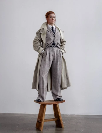 Woman in a gray suit and tan trench coat standing on a wooden stool in a minimalist setting, looking upward with red hair.