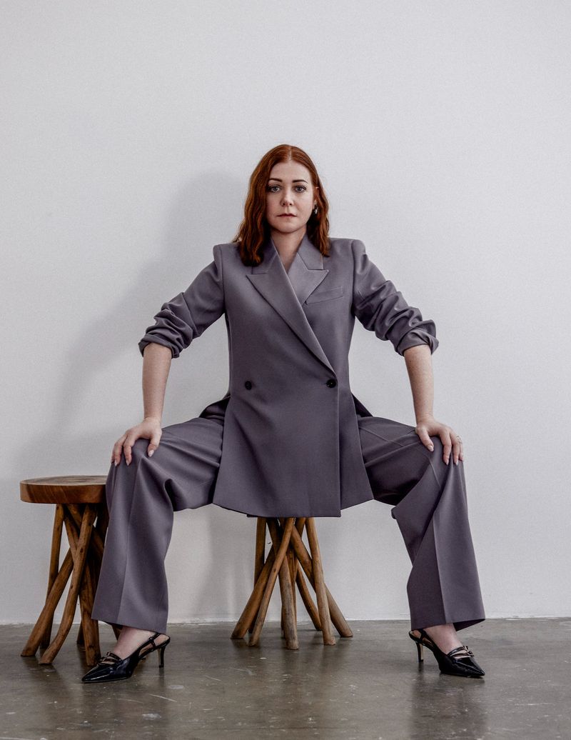 Adult woman with red hair sits on a wooden stool wearing a grey pantsuit with black heeled shoes in a light-filled indoor space.