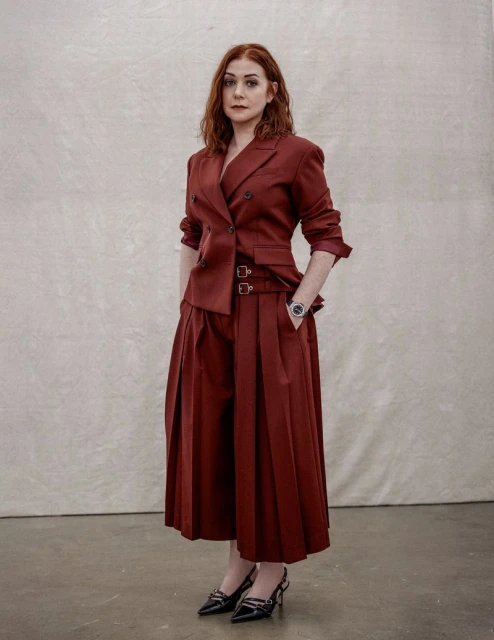 Portrait of a red-haired woman in a dark red blazer and pleated culottes, posing in front of a textured neutral background in a photo studio.
