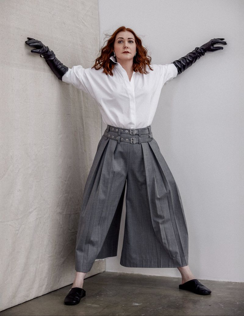 A red-haired woman in an oversized white shirt and gray culottes poses in a photo studio wearing long black leather gloves.